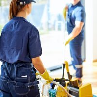 Back view on two janitors in blue working uniform clean up roon with panoramic window, wearing yellow rubber gloves